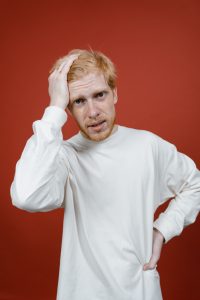 Portrait of a man in a white sweater showing confusion, hand on head against a red backdrop.